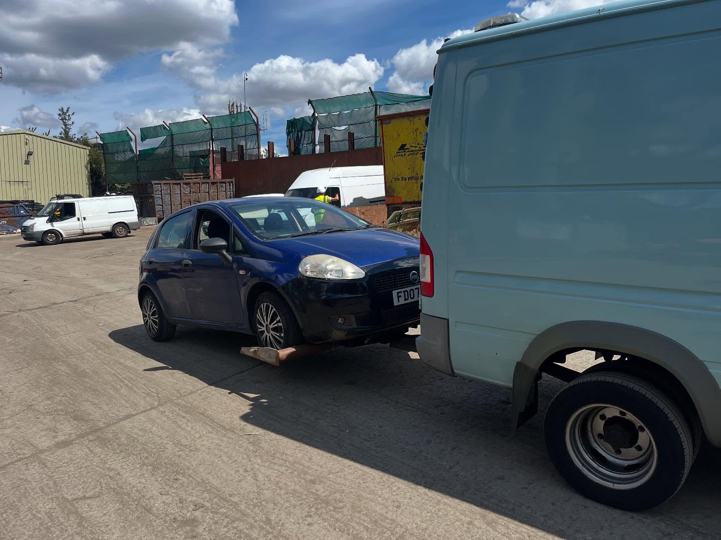 Dark Fiat Punto parked beside a van in the Leeds Scrap Cars yard under cloudy skies
